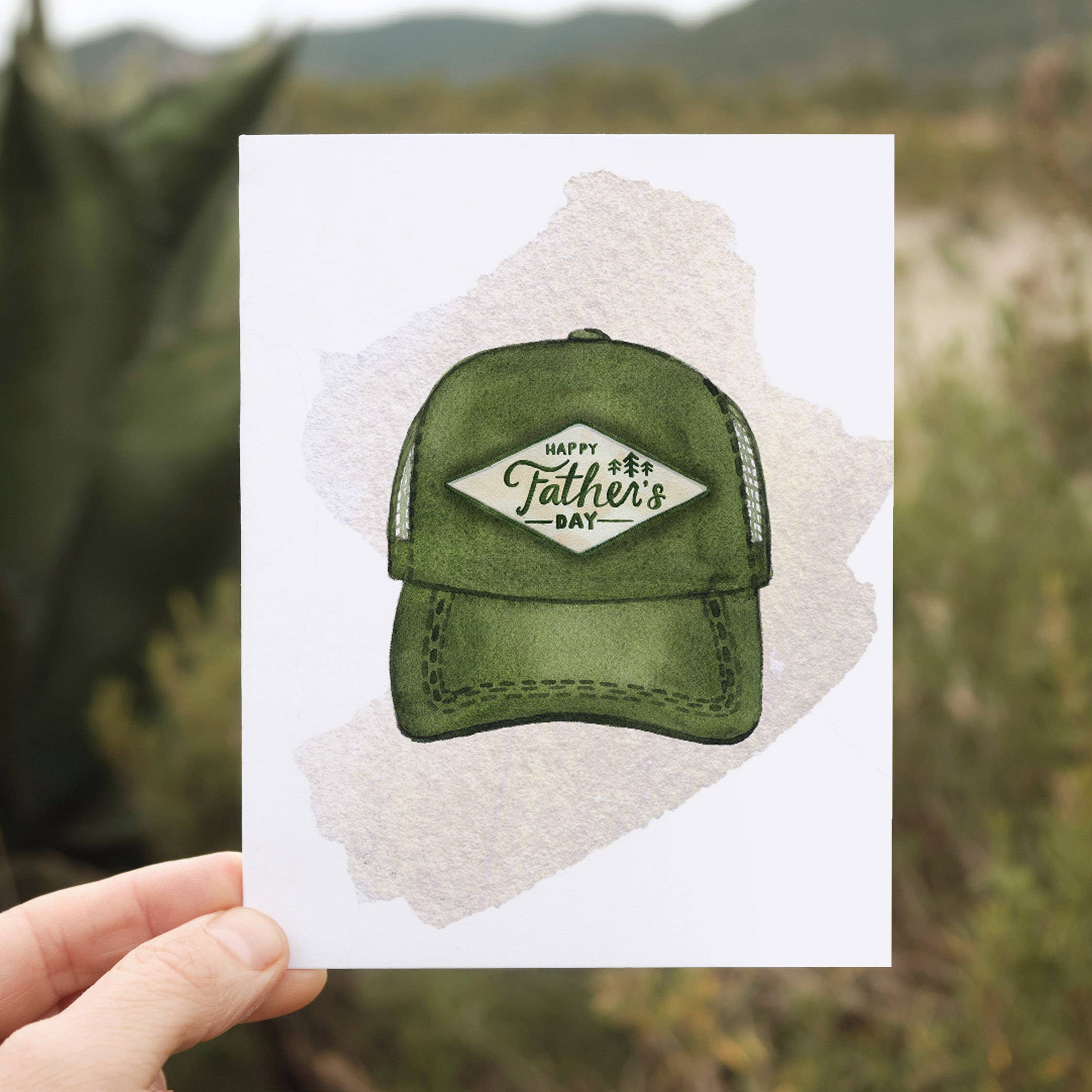 Hand holding a card with a green cap and 'Happy Father's Day' text against a natural background.