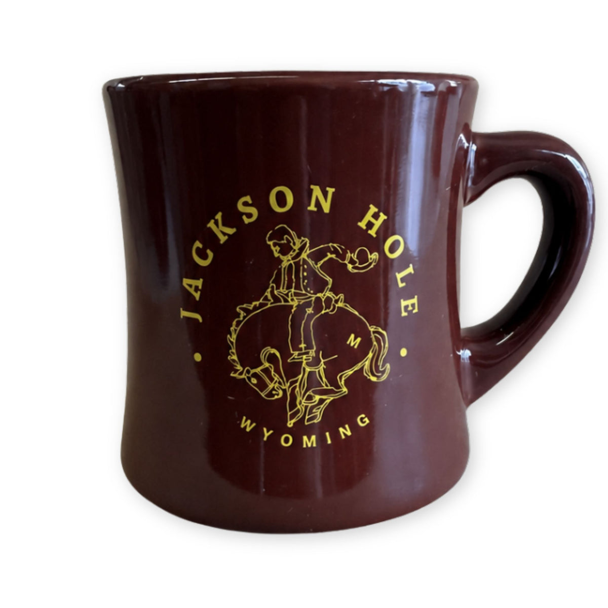 A glossy dark brown ceramic mug featuring a gold printed graphic of a cowboy on a bucking horse with the text 'JACKSON HOLE WYOMING' around it, shown on a plain white background.