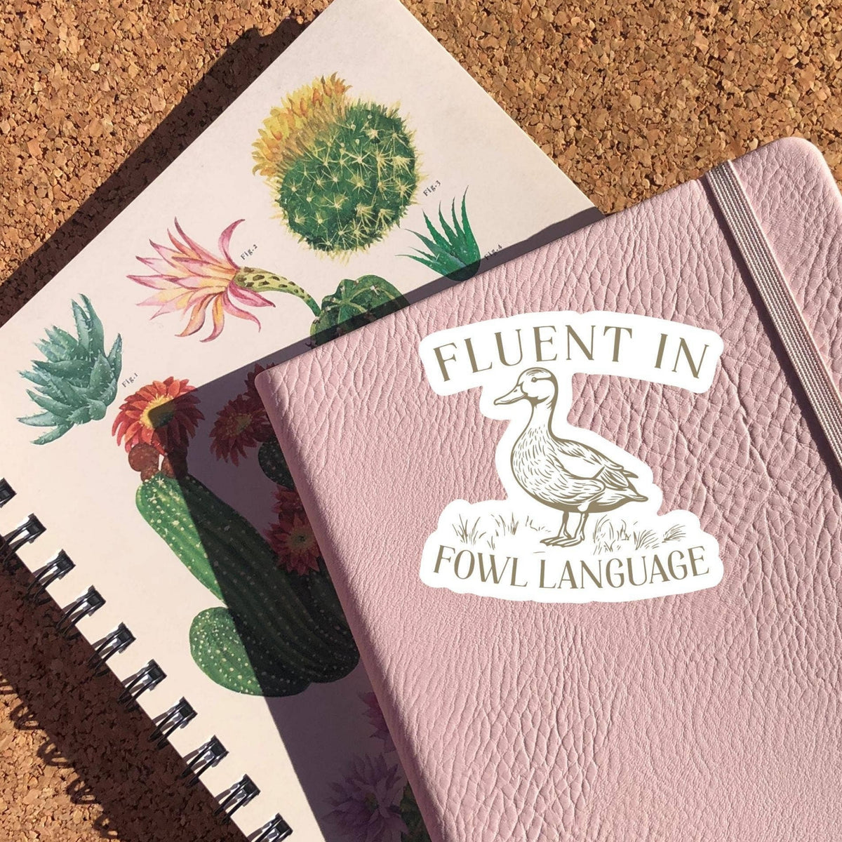 Fluent In Fowl Language Sticker on a pink notebook cover with a cactus print notebook beneath, glossy vinyl by Sticker Horse