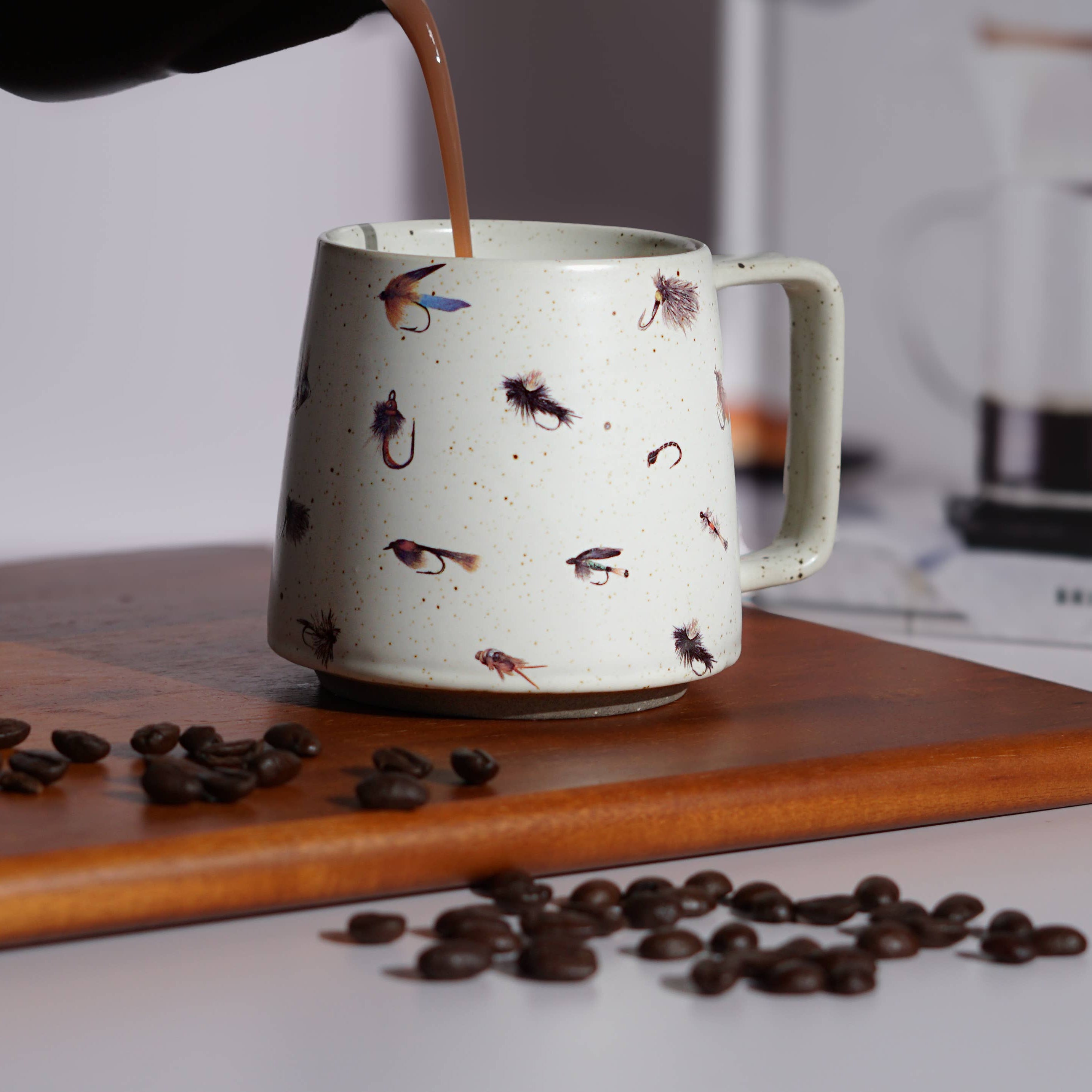 Coffee being poured into a white mug with bird patterns on a wooden board with scattered coffee beans.