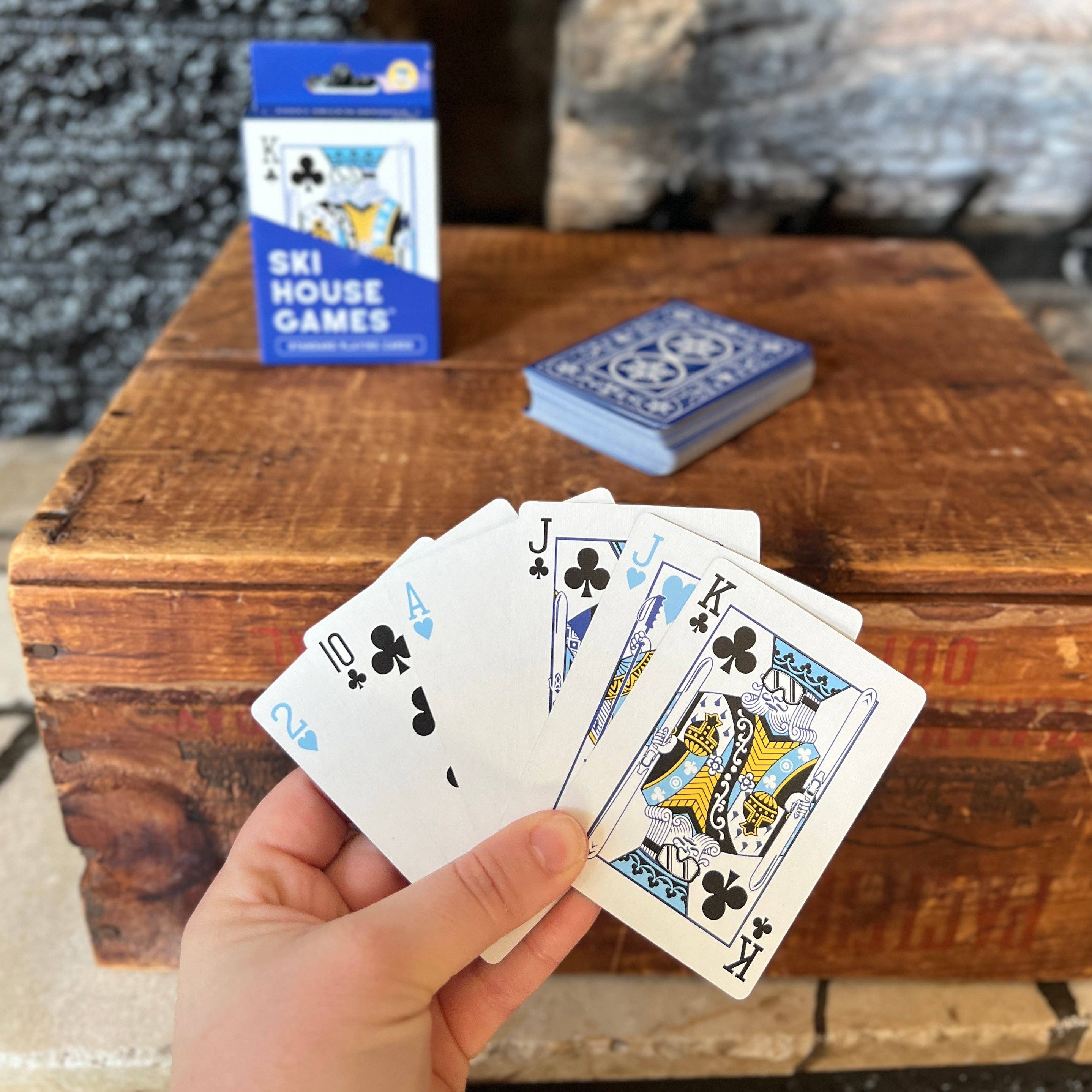 Hand holding playing cards with 'Ski House Games' packaging in the background on a wooden surface.