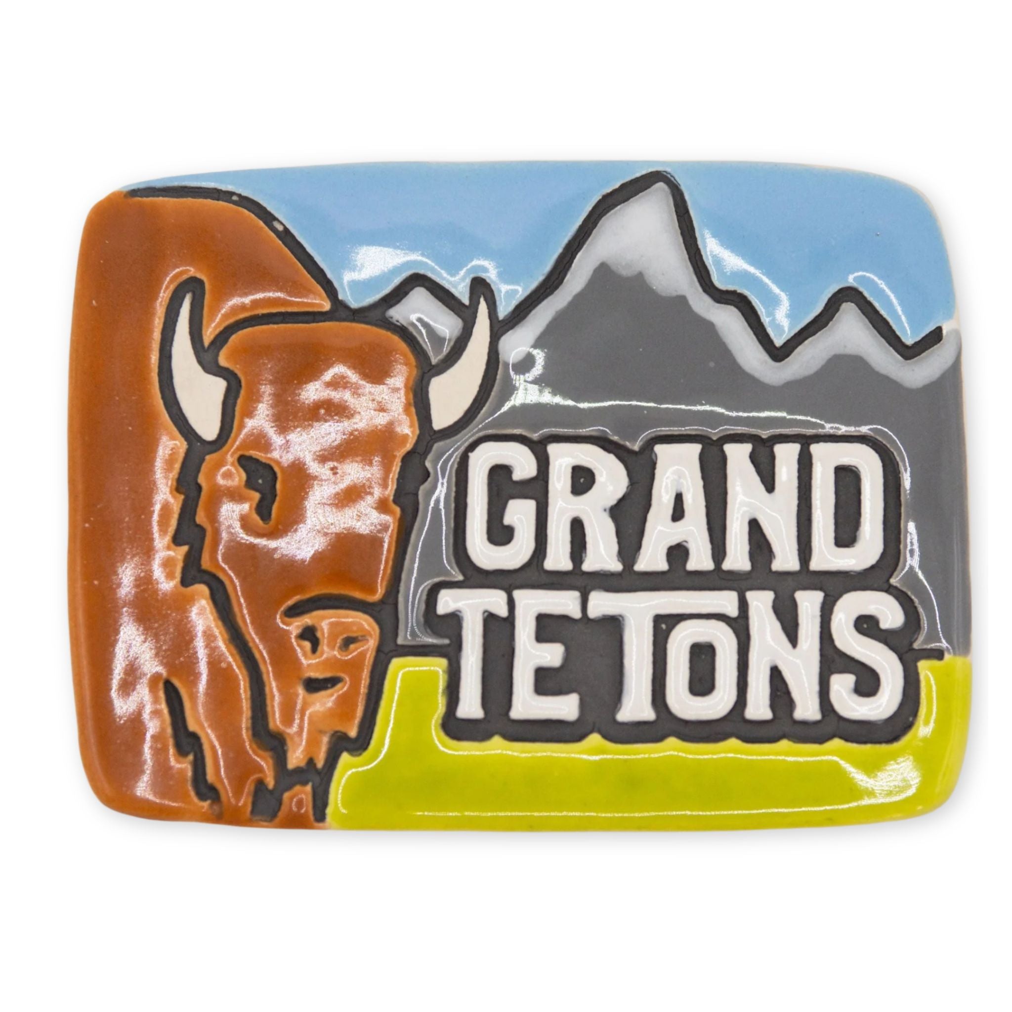 A rectangular ceramic magnet featuring a painted Grand Teton landscape with a brown bison on the left, gray mountains, a blue sky, and the words “GRAND TETONS” in large white letters with black outlines.