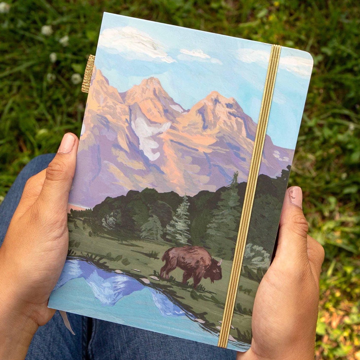 Person holding a notebook with a mountain and bear illustration on grass