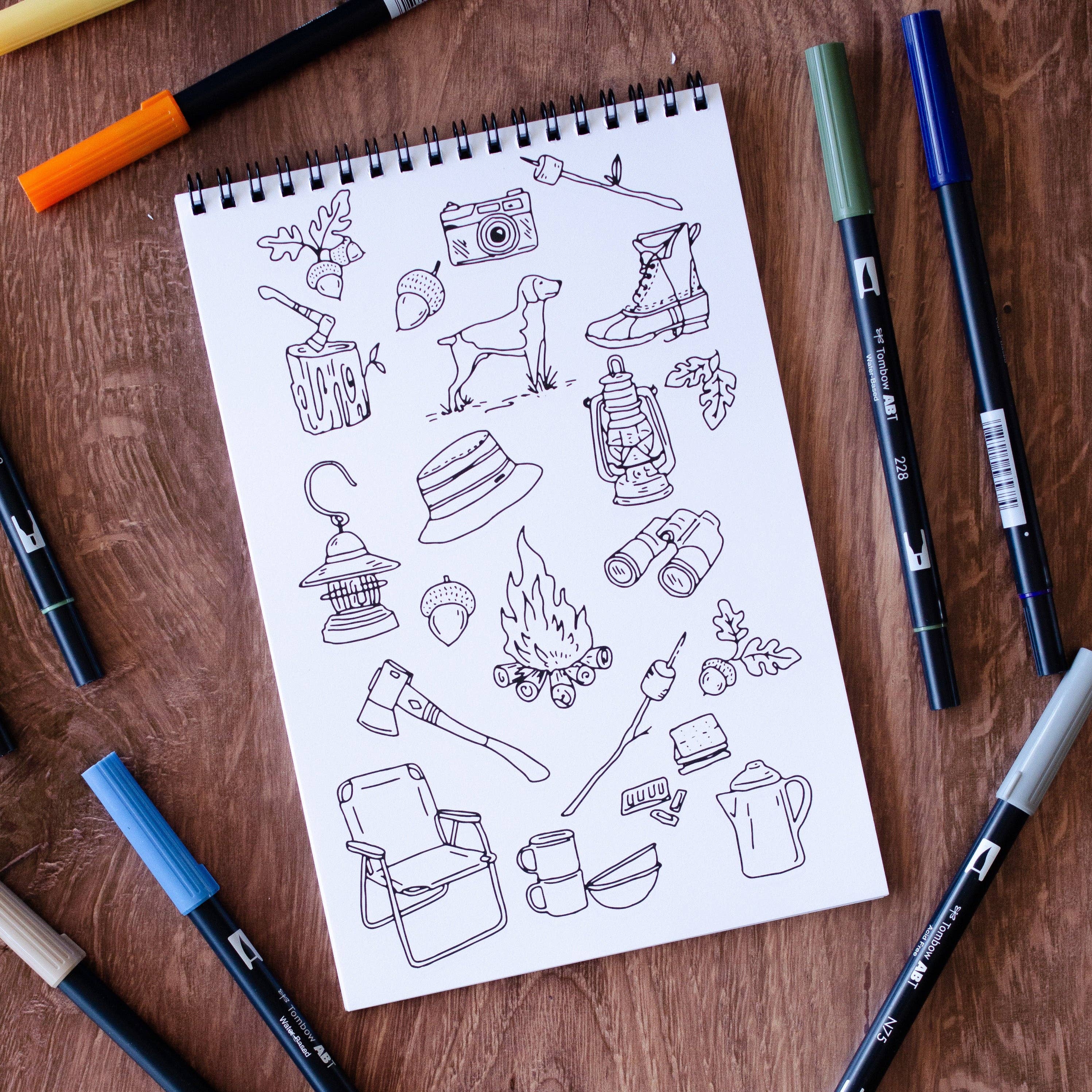 Notebook with camping-themed illustrations on a wooden surface with markers around.