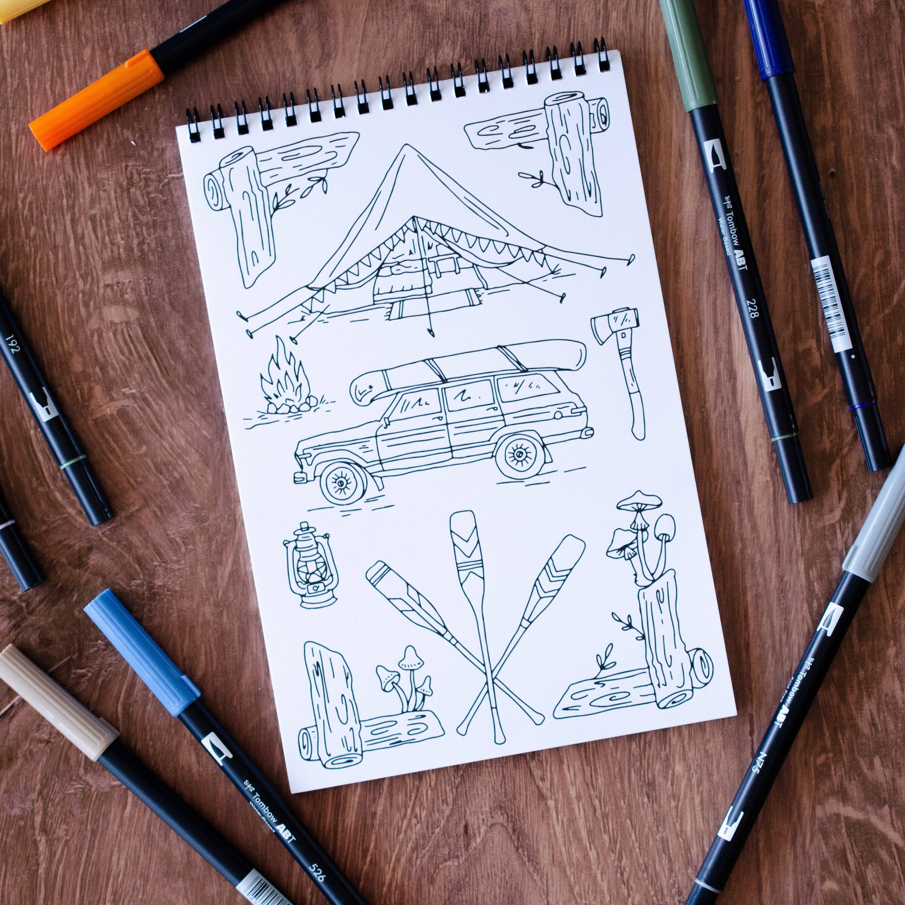 Notebook with camping-themed illustrations surrounded by markers on a wooden surface