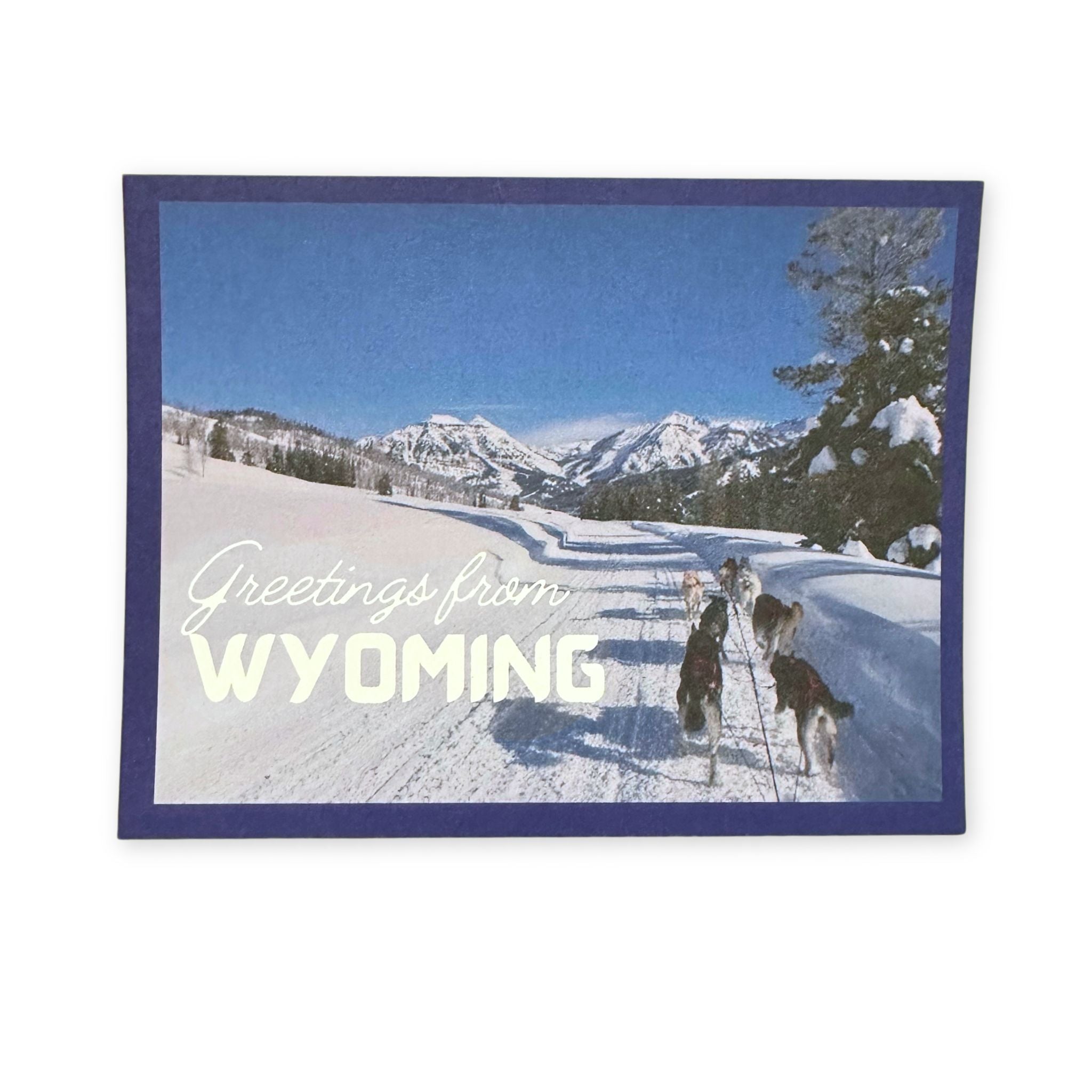 Dog sledding scene with mountains and 'Greetings from Wyoming' text on a snowy landscape.