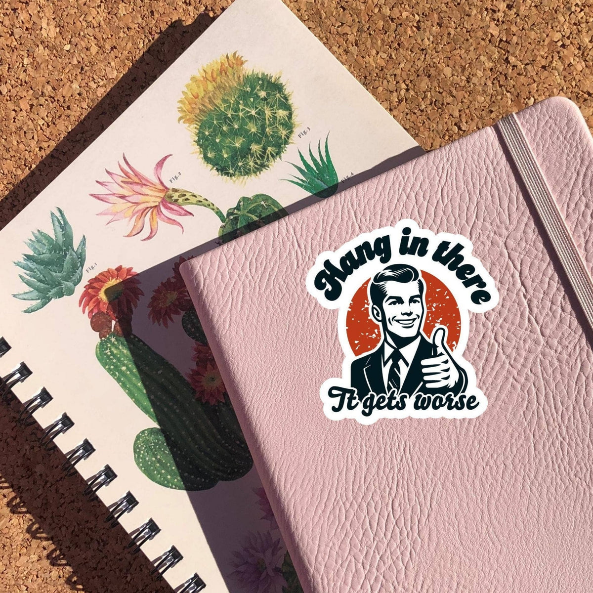 Hang In There It Gets Worse Sticker on a pink notebook cover with a cactus print notebook beneath