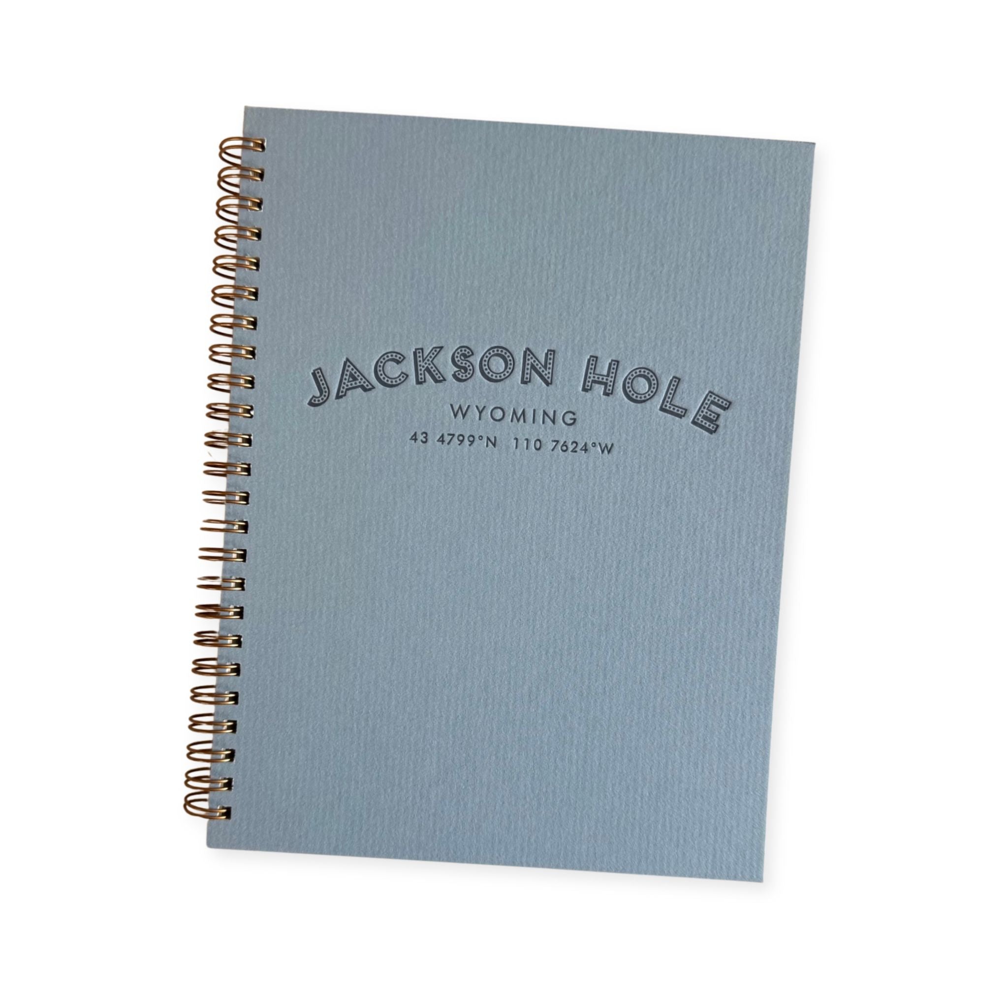 A light blue spiral-bound notebook with a textured cover, featuring the embossed text 'JACKSON HOLE WYOMING' and coordinates on the front. The notebook is shown at an angle with a gold-toned spiral binding on a clean white background.