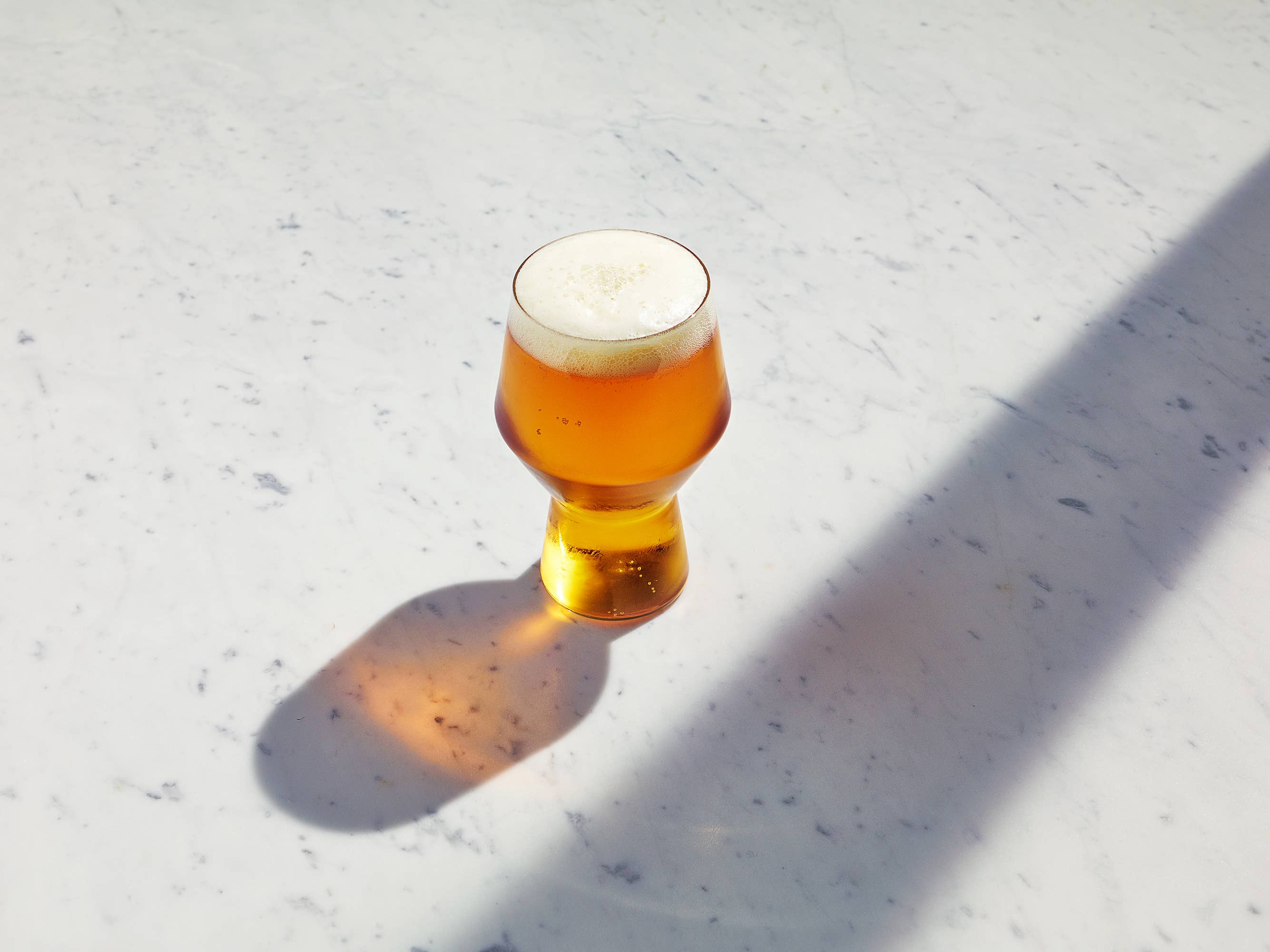 A small tulip-shaped glass filled with amber beer and topped with white foam, placed on a light marble surface with strong sunlight and shadows.