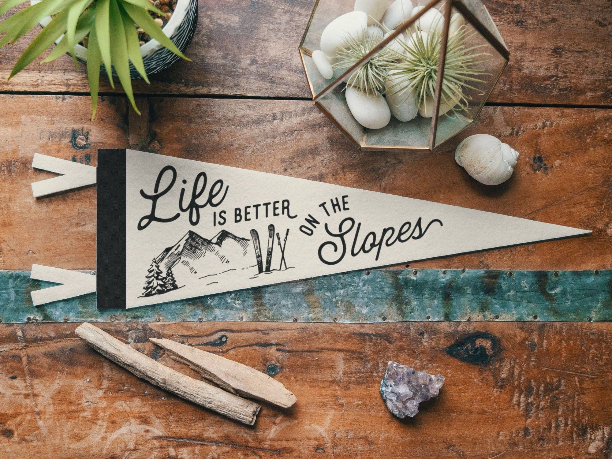 Decorative sign with 'Life is better on the slopes' text on a wooden surface with plants and stones.
