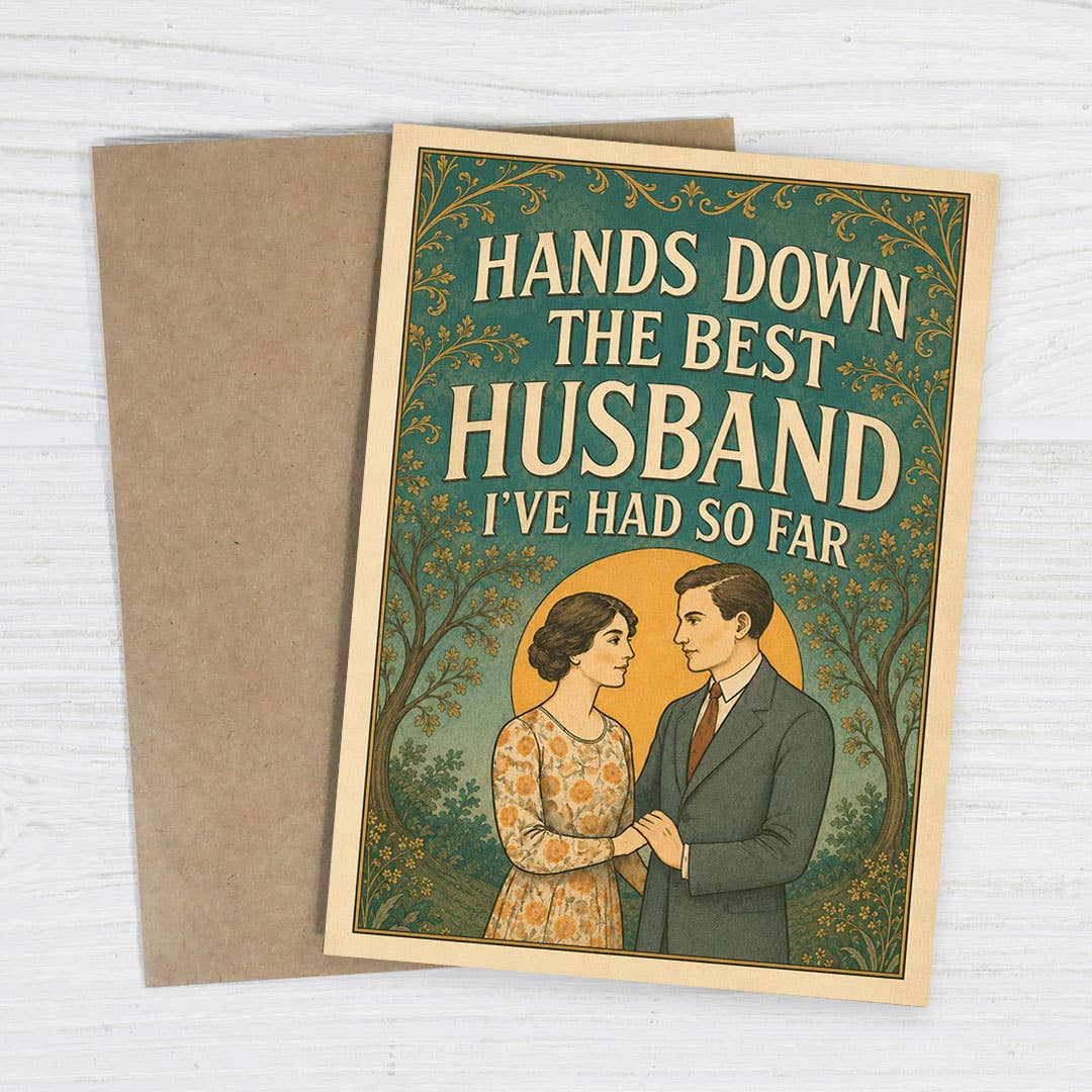 A rectangular greeting card with a kraft paper envelope, photographed on a white wood surface. The card has a vintage-style illustration of a couple under decorative trees, with the text 'HANDS DOWN THE BEST HUSBAND I'VE HAD SO FAR.