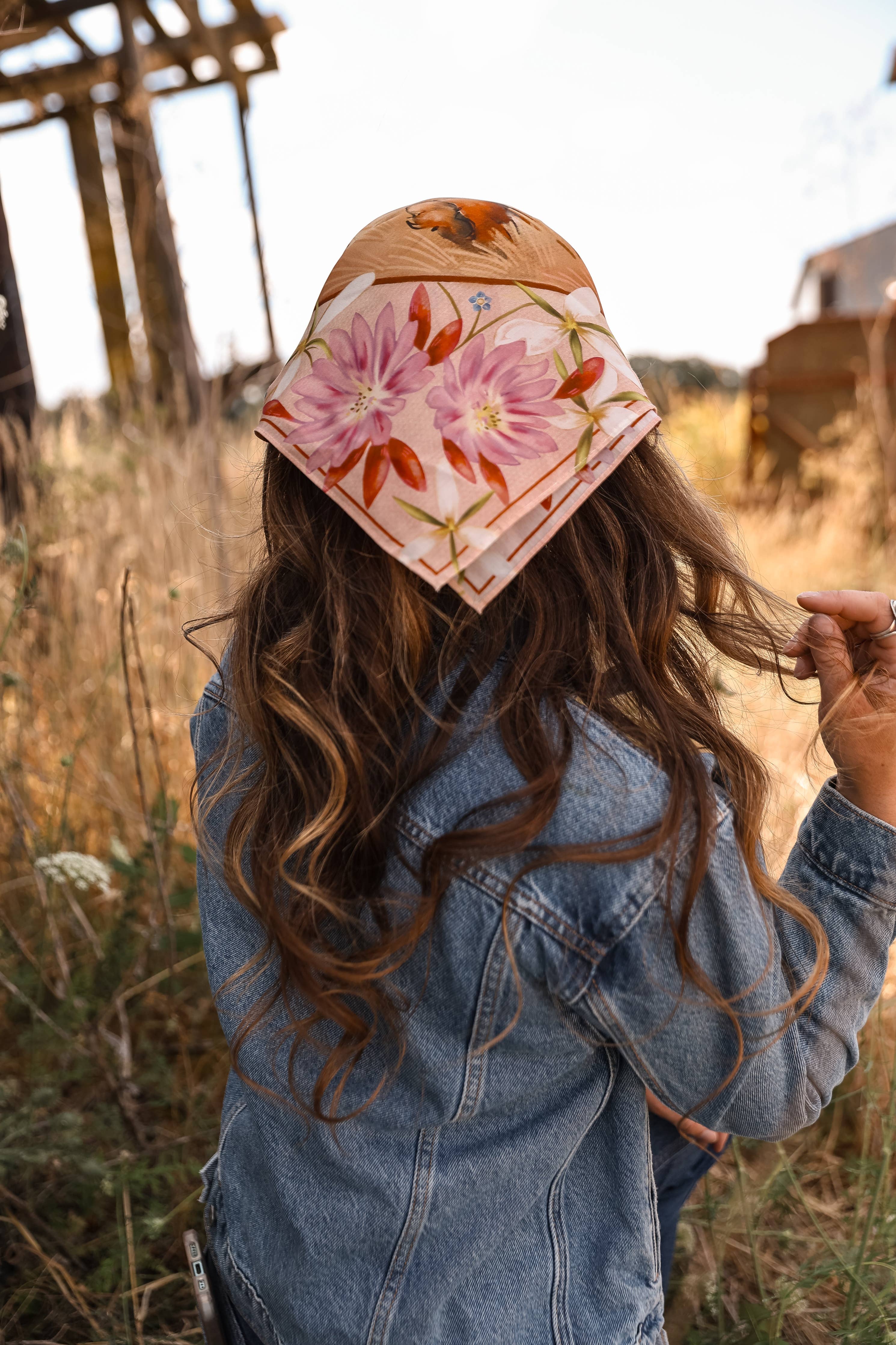 A person outdoors in a field, shown from behind wearing a floral bandana-style hat and a denim jacket.