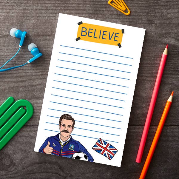 A white lined notepad with a yellow 'BELIEVE' banner and cartoon character illustration, styled on a dark wood surface with earbuds, paperclips, colored pencils, and a Union Jack sticker.