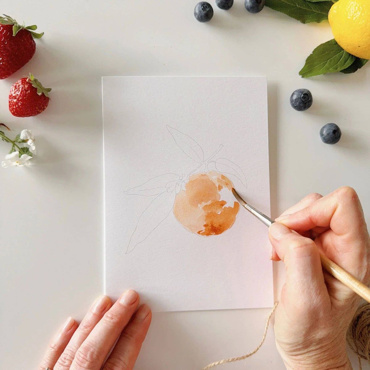 Person painting a watercolor of a orange on a white sheet with fruits and flowers around.