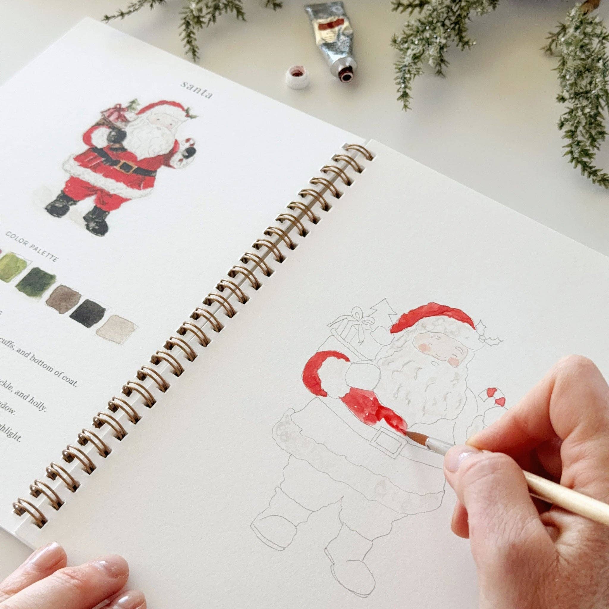 A hand uses a fine paintbrush to color a line drawing of Santa Claus in a spiral-bound sketchbook. The left page shows a finished Santa illustration with color swatches, and a tube of paint and greenery are visible on a light tabletop.