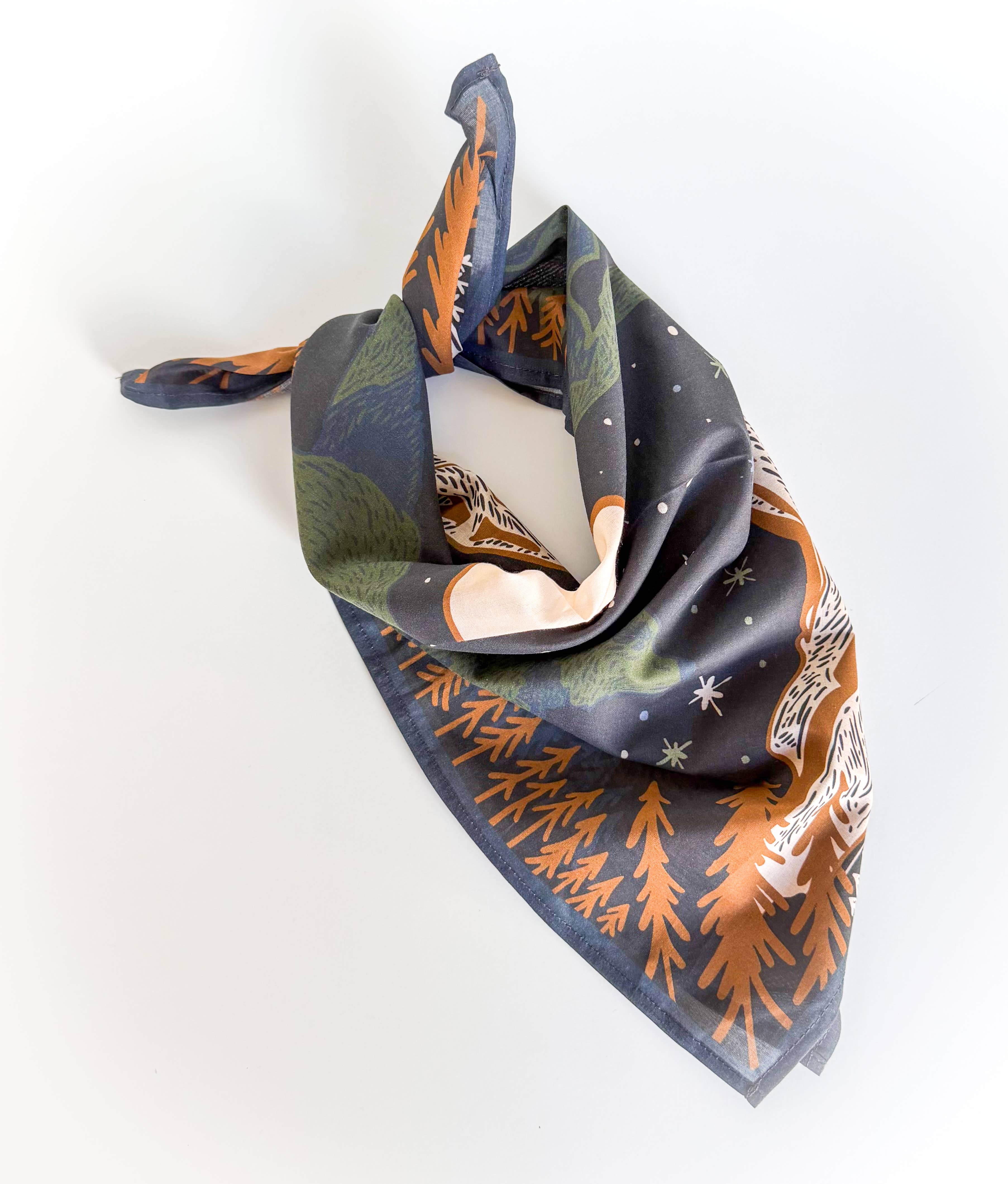 A square cotton scarf with a forest and winter pattern, featuring pine trees, snow, and stars in orange, green, and white on a dark navy background.
