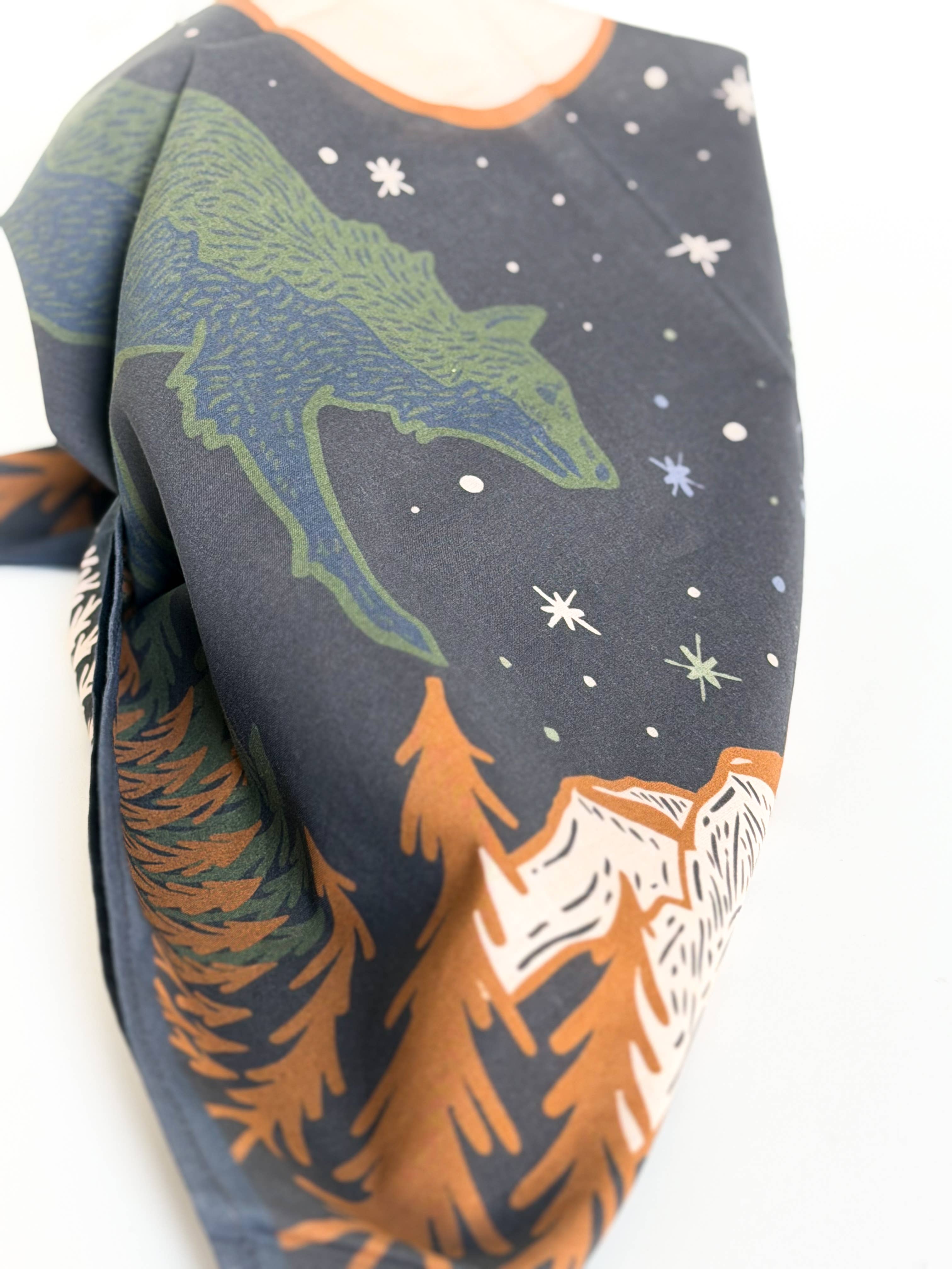 A close-up of a dark navy scarf featuring a green wolf, white stars, and orange and white pine trees and mountains, with a small portion of a beige garment visible at the top edge.