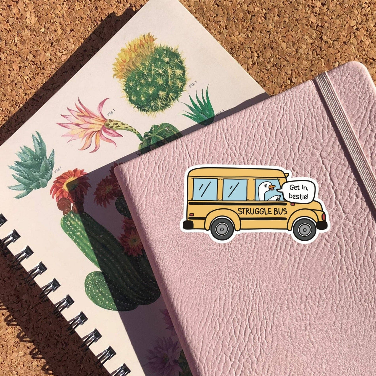 Struggle Bus Sticker on a pink notebook cover with a cactus print notebook beneath, glossy vinyl by Sticker Horse