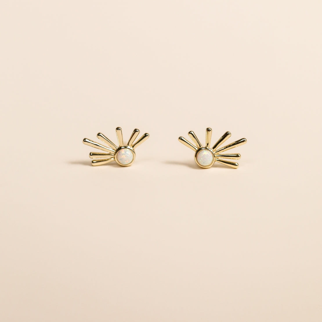 Gold sunburst earrings with a central white opal on a beige background