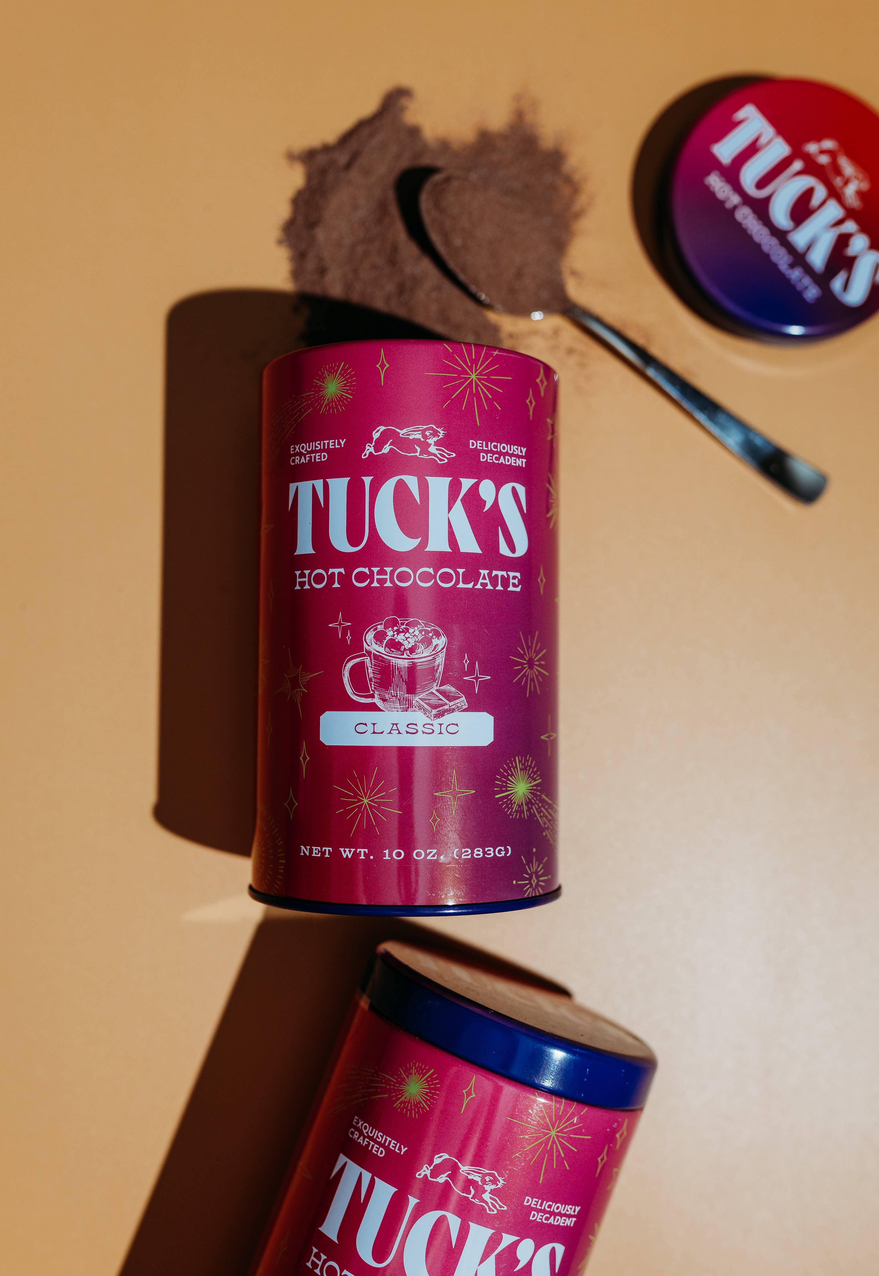 Tuck's Hot Chocolate canisters with a scoop of chocolate powder on a beige background