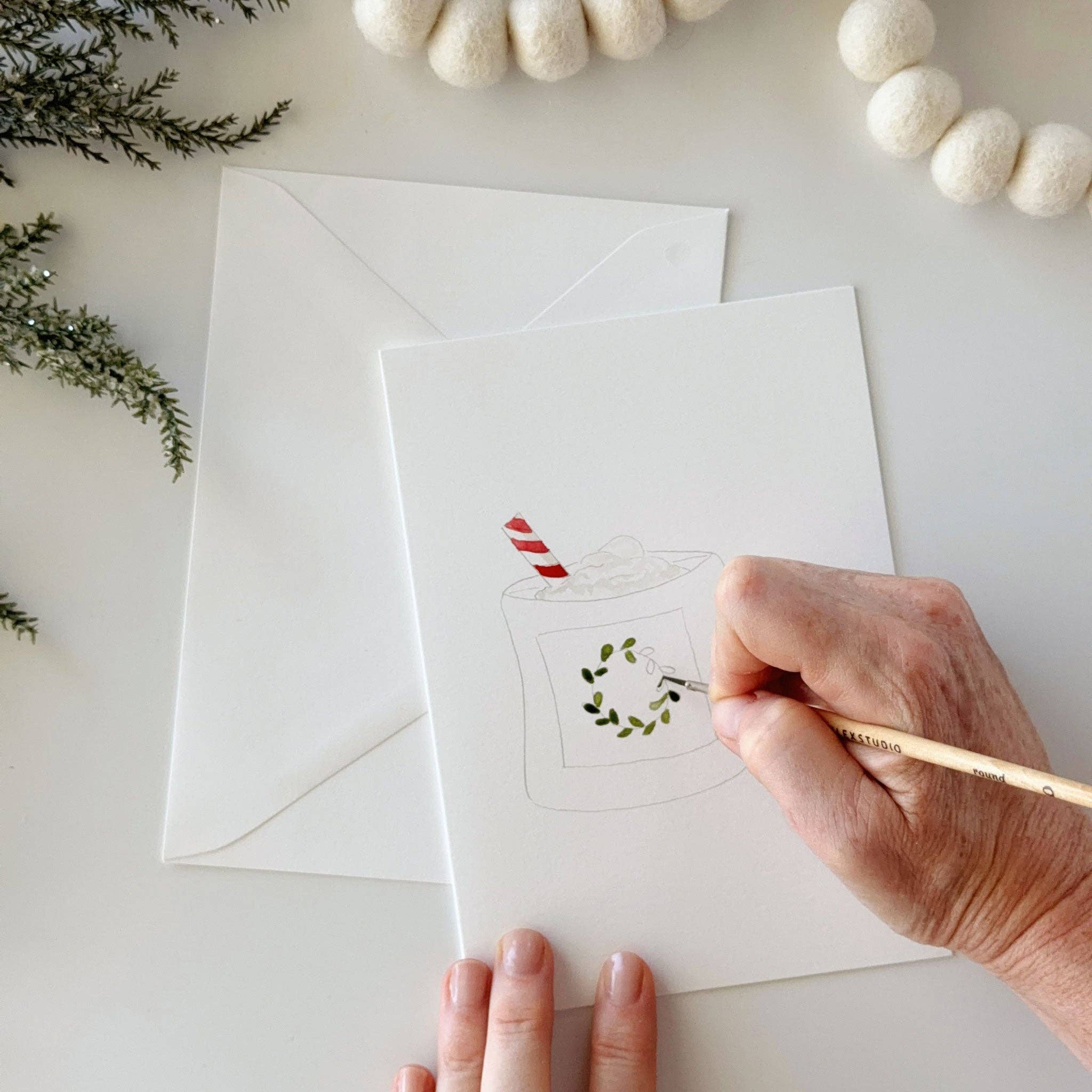 Painting on a white greeting card with a simple illustration of a hot chocolate cup and holiday greenery. A white envelope lies behind the card on a light surface, with decorative evergreen branches and white felt garlands around the scene.