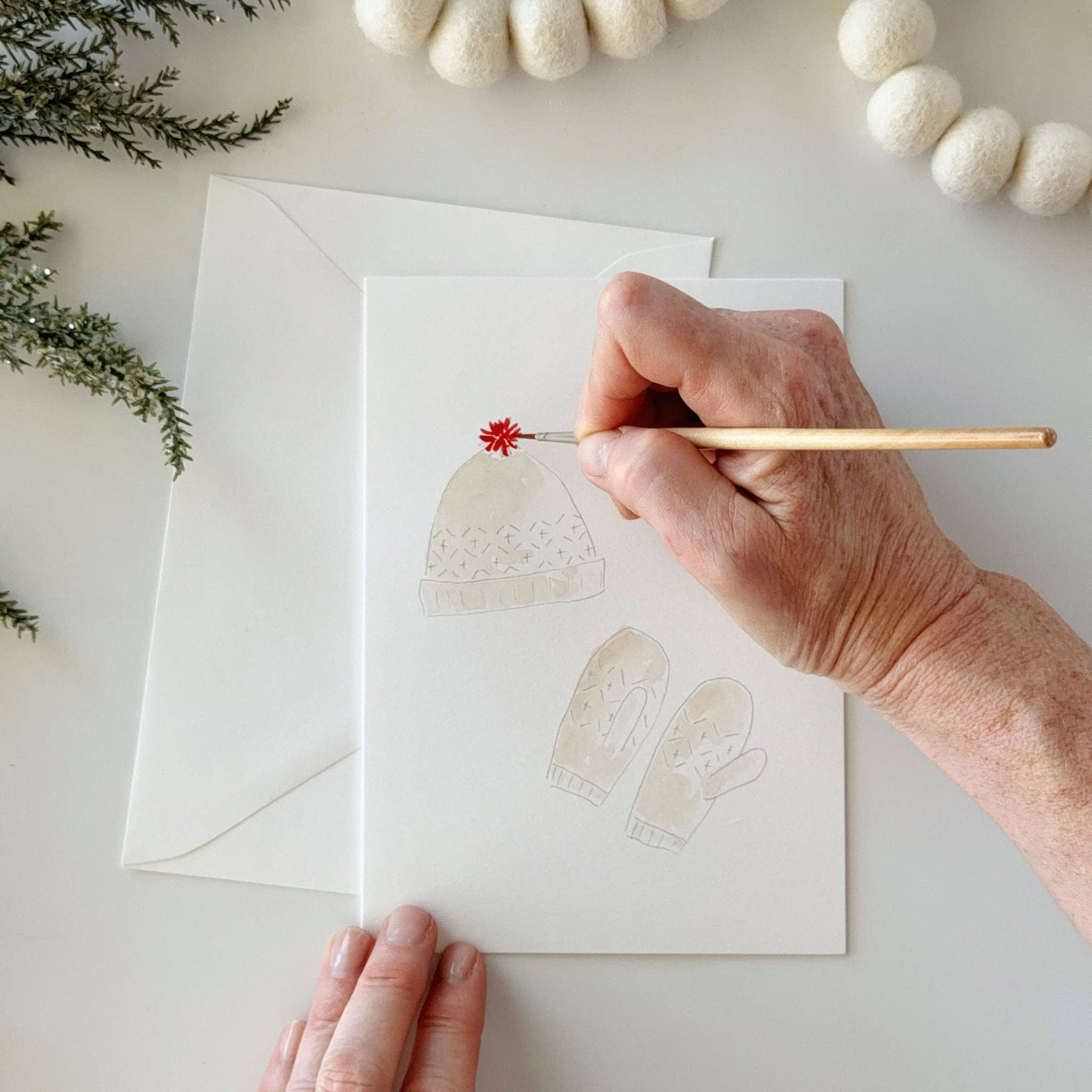 A close-up of a person painting on a white card with a light beige knit hat and mittens, with a matching white envelope behind it. The holiday scene is styled with white felt garlands and green pine branches on a light surface.