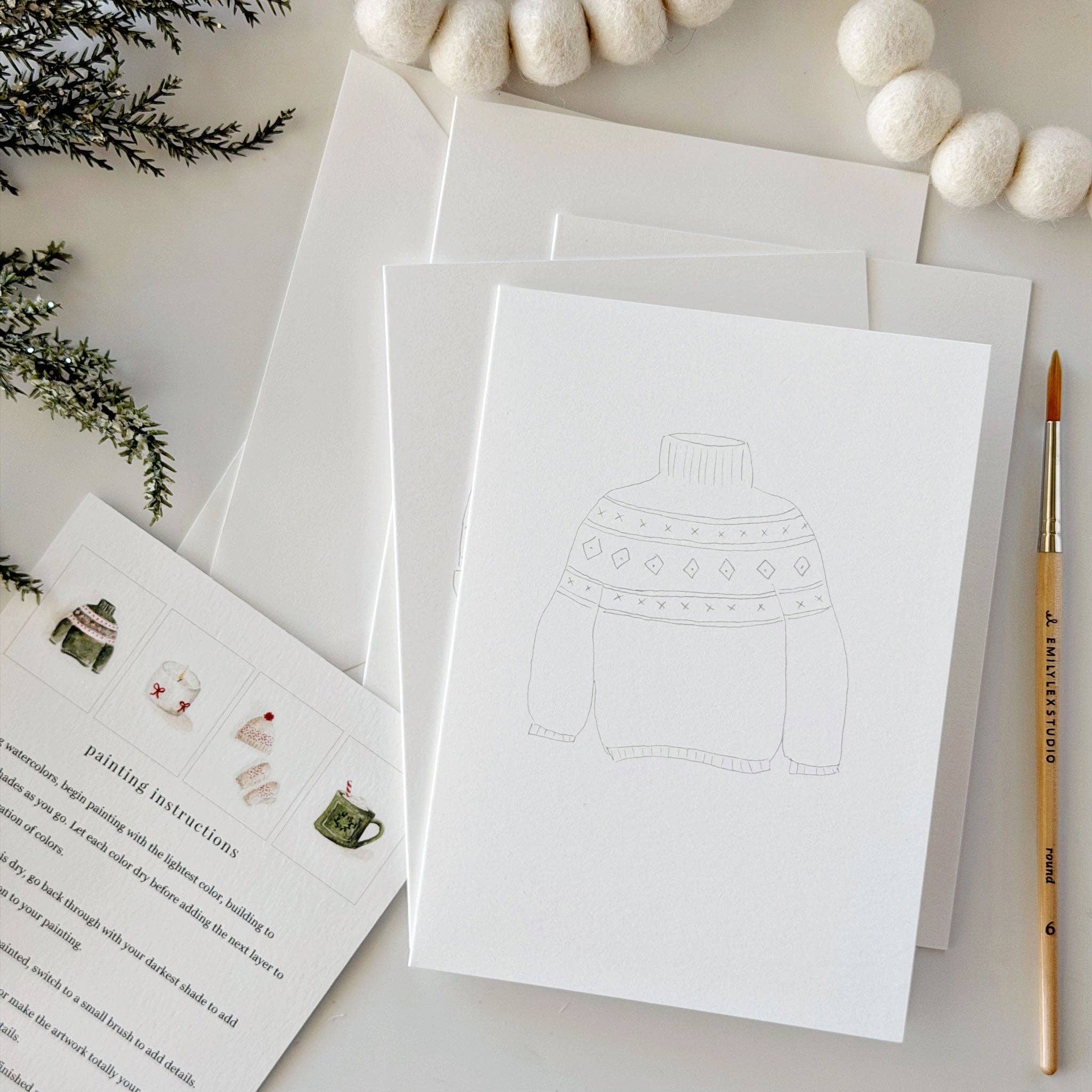 A flat-lay of a white art print featuring a faint line drawing of a sweater, shown with white envelopes, a printed painting instructions card, a paintbrush, and holiday greenery on a light background.