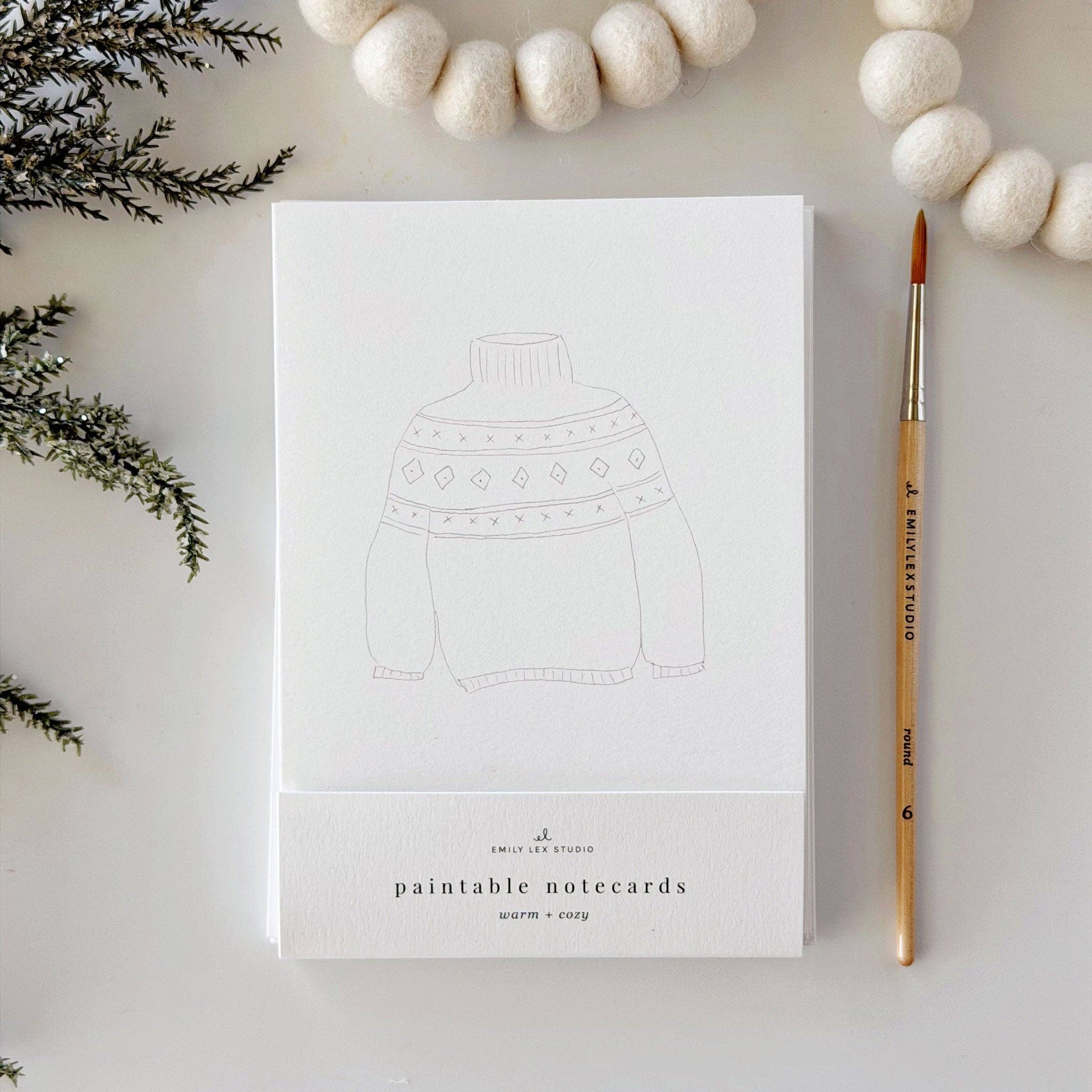 A white notepad with a faint sweater illustration and the text 'paintable notecards' and 'UNIQUELY STUDY' on the cover, styled on a light surface with a paintbrush and white felt balls.