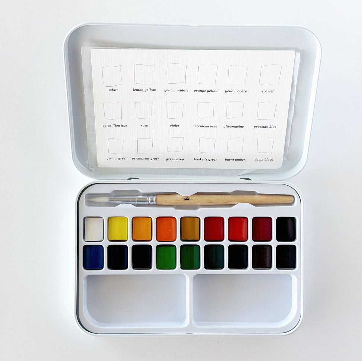 An open metal watercolor paint set with a white tray holding multiple color pans, a wooden-handled brush, and a sheet of color cards inside the lid.