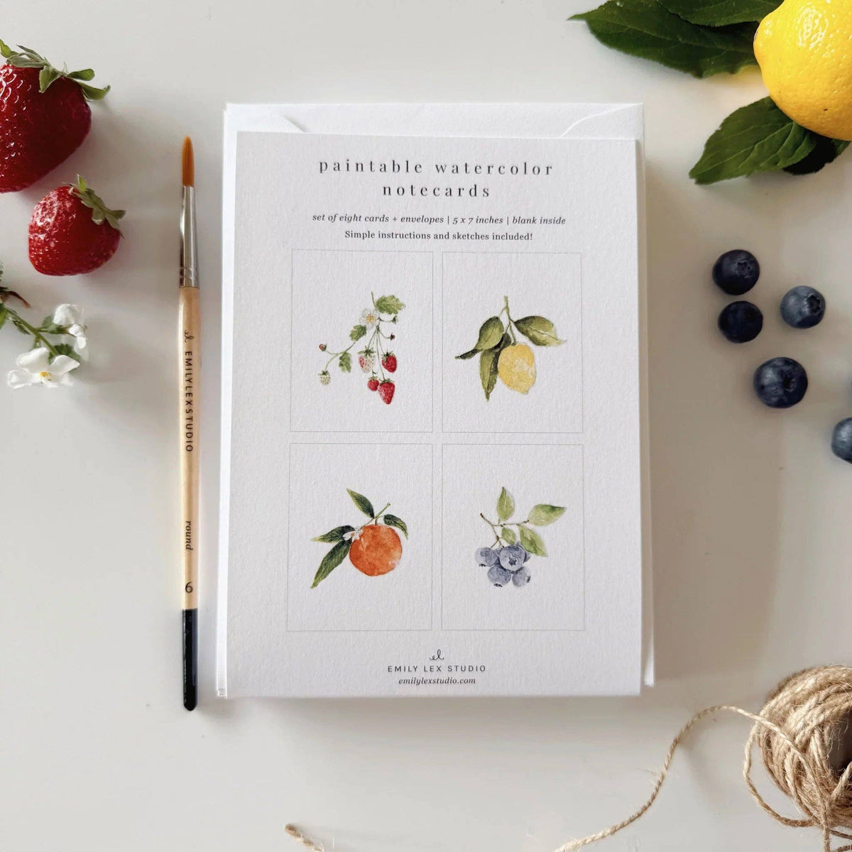 A flat-lay of Emily Lee Studio watercolor notecards on a white surface, surrounded by artistic props including a paintbrush, strawberries, blueberries, a lemon.