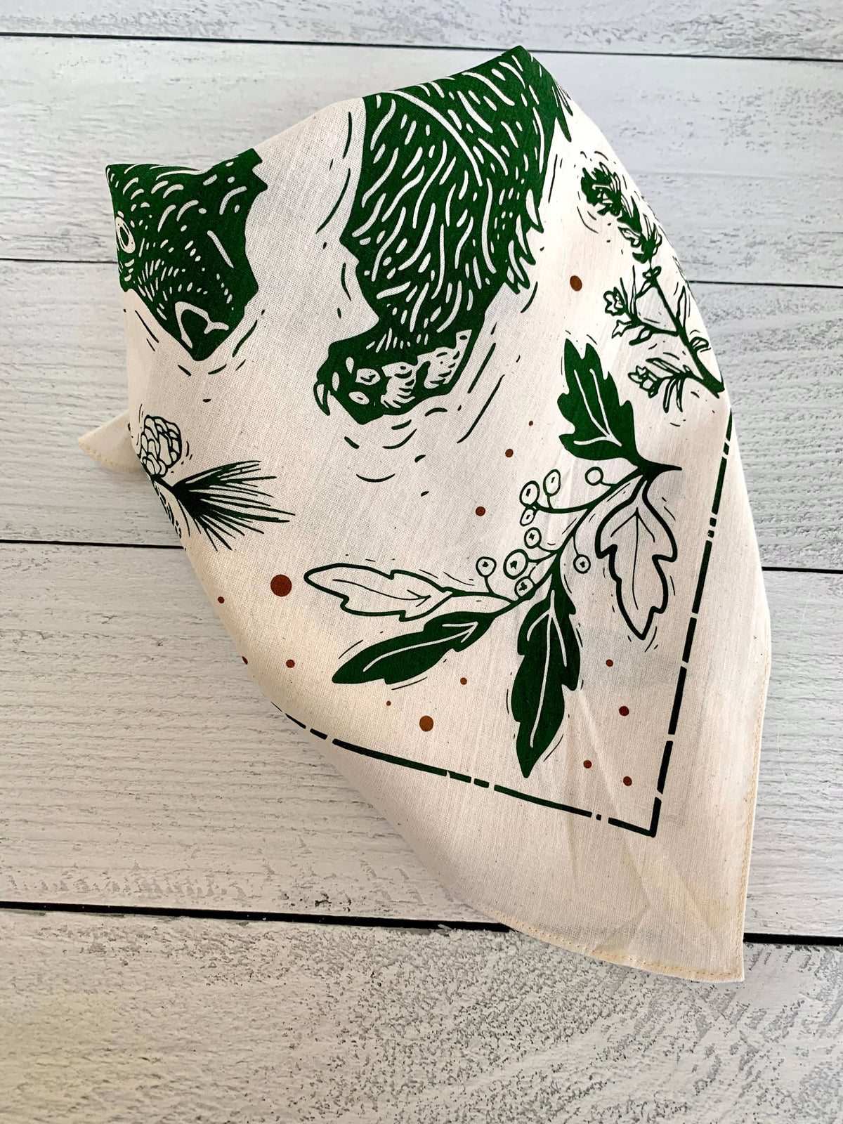 A square cream-colored bandana featuring a green and black woodland print of a bears and birds.