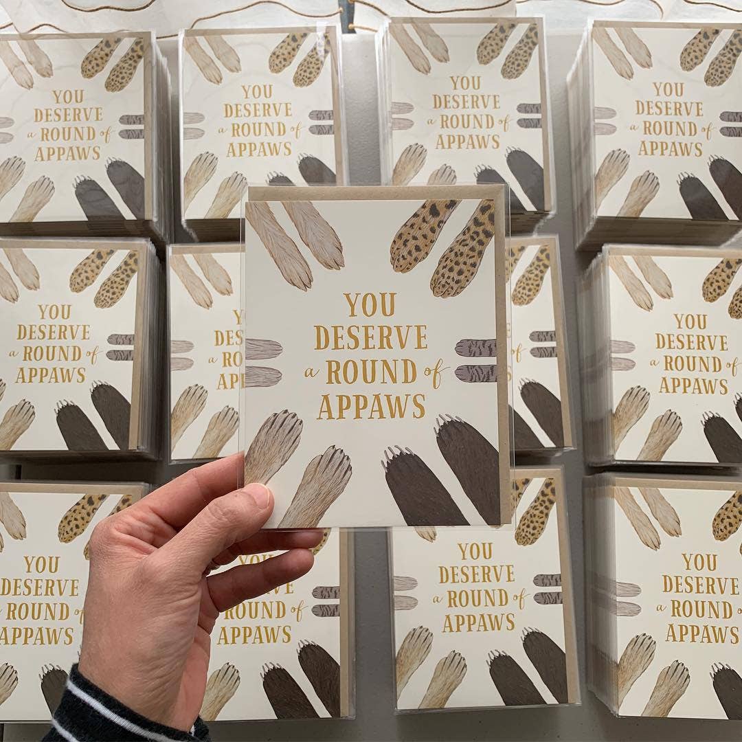 Hand holding a box with 'You Deserve Round of Appaws' text and paw prints on a reflective surface.