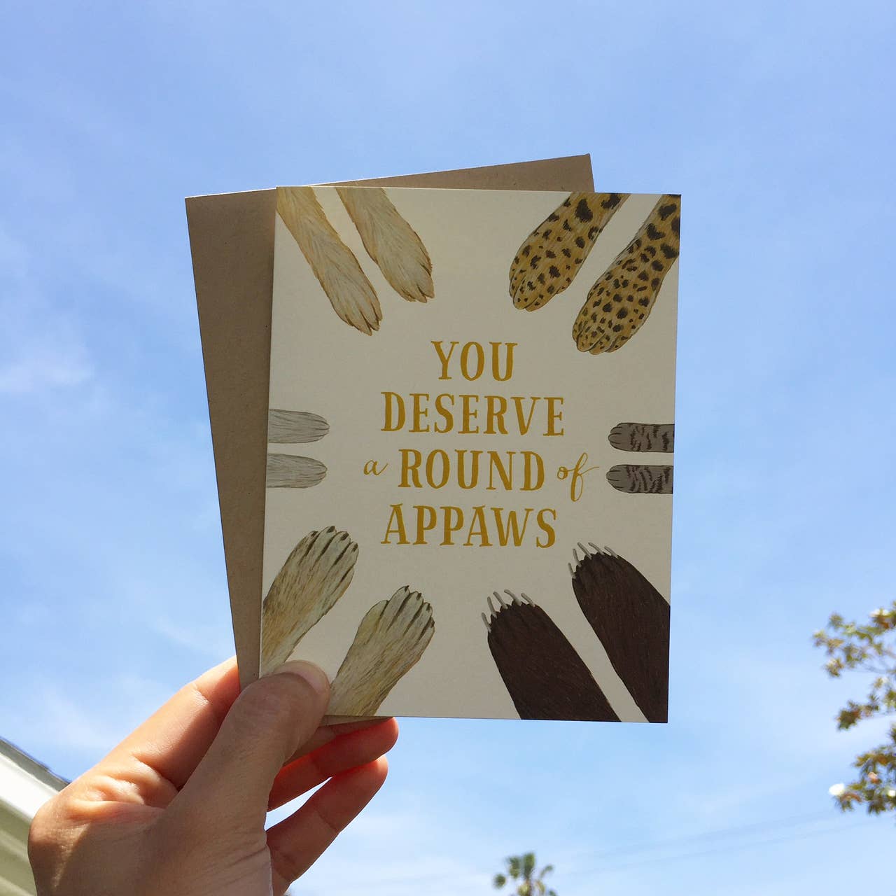Card with paw prints and text 'You Deserve a Round of Appaws' held against a blue sky.